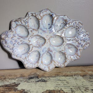 May include: A ceramic egg serving platter with a pearlescent glaze. The platter has 12 egg-shaped indentations and a scalloped edge. The color is primarily white with hints of blue and brown.