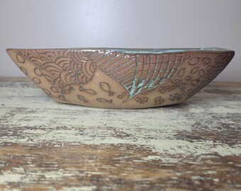 May include: A shallow, hand-crafted ceramic bowl with a tan, unglazed exterior featuring etched patterns of fish, leaves, and geometric lines. The interior rim is accented with a vibrant turquoise glaze that contrasts with the earthy clay tone.