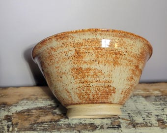 May include: A handmade ceramic bowl with a speckled, earthy design. The bowl features a light beige base color with brown speckles and a slightly flared rim. The bowl sits on a small, circular base, showcasing a rustic, artisanal aesthetic.
