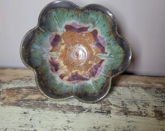 May include: A flower-shaped ceramic bowl featuring a reactive glaze with a central speckled golden-brown base, transitioning into deep purple accents and a vibrant green rim. The bowl has a smooth, glossy finish and a scalloped edge design.