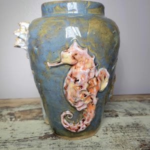 May include: A blue ceramic vase with a seahorse design. The vase has a textured surface and is adorned with a raised, coral-colored seahorse. The vase has a wide base and a narrower neck. The seahorse is detailed with brown spots.