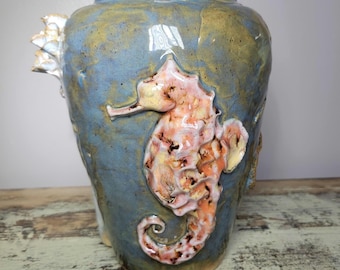 May include: A blue ceramic vase with a seahorse design. The vase has a textured surface and is adorned with a raised, coral-colored seahorse. The vase has a wide base and a narrower neck. The seahorse is detailed with brown spots.