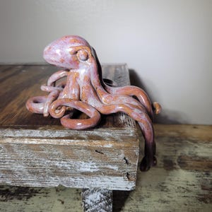 Könnte beinhalten: Eine Keramik-Oktopus-Figur mit glänzender Oberfläche in Rosa-, Lila- und Orangetönen. Die Skulptur hat einen abgerundeten Kopf mit sichtbaren Augenvertiefungen und mehrere geschwungene Tentakel, die nach außen ragen und über den Rand hängen.