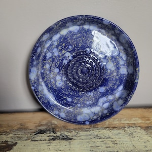 May include: A handmade ceramic grater plate with a deep blue glaze and speckled white and yellow accents. The center features a textured grating surface. The plate is round and sits on a wooden surface.