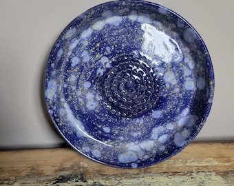 May include: A handmade ceramic grater plate with a deep blue glaze and speckled white and yellow accents. The center features a textured grating surface. The plate is round and sits on a wooden surface.