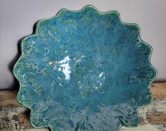 May include: A turquoise ceramic bowl with a scalloped edge. The bowl features a detailed floral pattern and a glossy finish. The rim and details are a darker shade of green. The bowl sits on a distressed wooden surface.