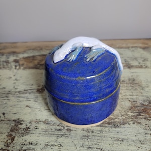 以下が含まれることがあります： コバルトブルーの釉薬を施した手作りの陶器製の小物入れ。白いトカゲの彫刻が蓋の上にあります。箱は丸く、蓋がぴったりと収まります。トカゲは青い足と白い体を持っています。