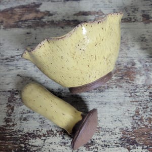 May include: A yellow ceramic mortar and pestle set. The mortar has a ruffled edge and a speckled finish. The pestle is cylindrical with a flat, brown base. Both pieces have a brown rim and base.