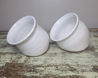 May include: Two off-white ceramic planters with speckled detailing. The planters have a rounded shape with a tilted opening. They are set on a distressed wooden surface, with a neutral background.