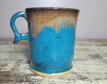 May include: A handmade ceramic mug with a unique design. The mug features a turquoise blue glaze that transitions to a tan and brown rim. The handle is a matching turquoise blue. The mug is approximately 4 inches tall.