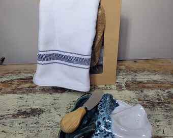 May include: A cheese board set with a white and blue ceramic dish, a wooden-handled knife, and a white towel with blue stripes. The set is presented with a brown gift bag.