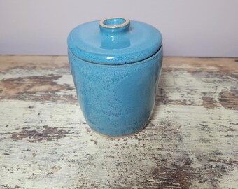 May include: A turquoise ceramic butter crock with a matching lid featuring a small knob handle. The glaze has a glossy finish with subtle variations in color intensity across the rounded body, showcasing a smooth texture and a clean, simple design.