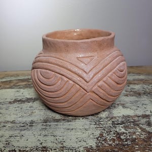 May include: A peach-colored ceramic pot with a rounded body and a slightly flared rim. The pot features carved geometric patterns, including curved lines and a triangle design. The texture of the pot is visible, adding to its handcrafted appearance.