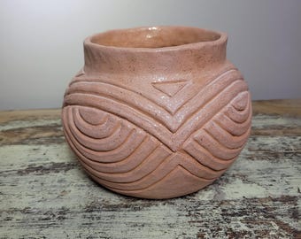 May include: A peach-colored ceramic pot with a rounded body and a slightly flared rim. The pot features carved geometric patterns, including curved lines and a triangle design. The texture of the pot is visible, adding to its handcrafted appearance.