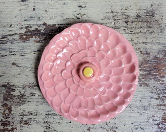 May include: A pink ceramic dish with a flower design. The dish has a central yellow accent and petal-like details. The background is a distressed wooden surface.