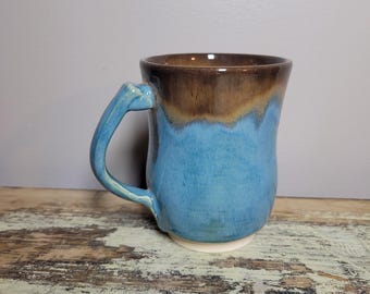 May include: A handmade ceramic mug with a blue and brown glaze. The mug has a rounded body, a sturdy handle, and a white base. The interior is brown, and the exterior is a vibrant blue with a brown rim. This unique mug is perfect for coffee or tea.