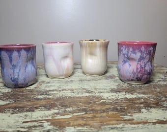 May include: Four ceramic cups with irregular, dimpled shapes. The first cup has a blue and purple glaze, the second is pale pink and white, the third is a creamy beige, and the fourth features a vibrant pink and purple mottled glaze pattern.