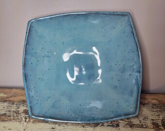 May include: A square ceramic bowl with a light blue glaze and dark blue speckles throughout. The bowl features slightly curved sides and a smooth, glossy finish, with a darker blue rim that defines the square shape of the piece.