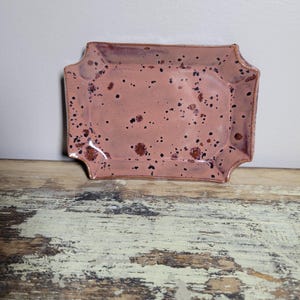 May include: A small, rectangular ceramic dish with a pink glaze and brown speckles. The dish has a slightly scalloped edge.
