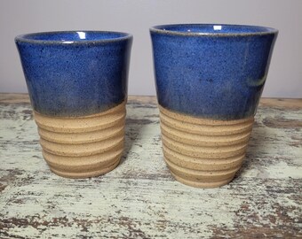 May include: Two ceramic tumblers featuring a smooth, deep blue glazed upper section and a textured, unglazed tan base with horizontal ridges. The vessels have a cylindrical shape with a slightly wider rim and a speckled finish on the lower portion.