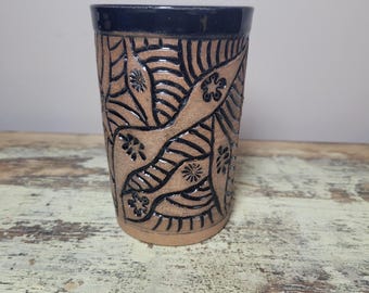 May include: A cylindrical ceramic tumbler featuring a tan, speckled clay body with a dark navy blue rim. The exterior is decorated with deep, carved black lines forming abstract leaf-like patterns and small flower shapes across the surface.