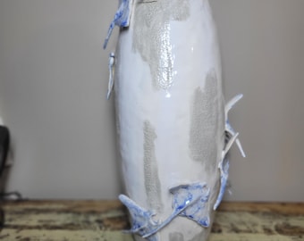 May include: A tall, cylindrical ceramic vase with a white glazed finish and patches of textured, matte gray clay. The surface is adorned with several three-dimensional, hand-sculpted stingray figures featuring blue and white mottled patterns.