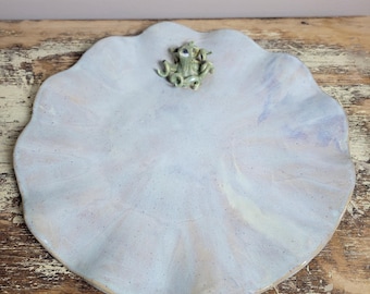 May include: A ceramic plate with a light blue and pinkish hue, shaped like a lily pad. A small, green frog figurine is centered on the plate. The plate has a wavy edge and a speckled texture, perfect for serving food.