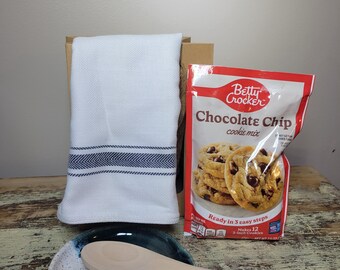 May include: A white kitchen towel with a blue chevron stripe, a ceramic spoon rest with a blue and white design, a wooden spoon, and a Betty Crocker Chocolate Chip cookie mix package. The package says "Ready in 3 easy steps".
