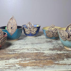 May include: A collection of handmade ceramic bowls in various colors, including red, blue, and yellow. Each bowl features a decorative leaf design and a leaf-shaped handle. The bowls are displayed on a distressed wooden surface.