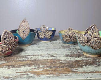 May include: A collection of handmade ceramic bowls in various colors, including red, blue, and yellow. Each bowl features a decorative leaf design and a leaf-shaped handle. The bowls are displayed on a distressed wooden surface.
