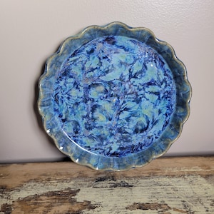 May include: A round, ceramic pie plate with a wavy edge. The plate features a vibrant blue glaze with a marbled pattern of lighter blues and hints of gold. The rim has a darker blue-green hue.