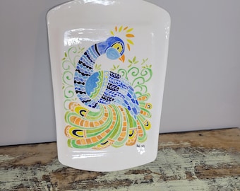 May include: White ceramic decorative tray with a hand-painted peacock design. The peacock features blue, green, yellow, and orange feathers. The tray has a curved top and a slightly curved bottom.