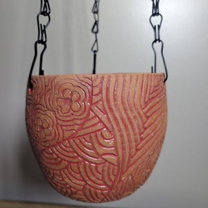 Op de afbeelding: Een hangende terracotta plantenbak met een gedetailleerd bloemen- en golfpatroon dat in het oppervlak is gesneden. De plantenbak wordt opgehangen door zwarte metalen kettingen, waardoor een decoratief en functioneel stuk ontstaat voor het tentoonstellen van planten. De kleur is een warme, aardse tint.