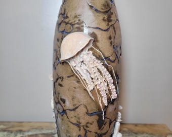 May include: A tall, cylindrical ceramic vase with a textured, mottled brown surface featuring dark, crackled lines. A sculpted, three-dimensional light pink jellyfish with long, trailing tentacles is attached to the front of the vase.