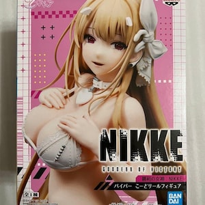 May include: A boxed anime figure of a female character with long blonde hair, wearing a white bra. The box is pink and white with the text "NIKKE GODDESS OF VICTORY" and Japanese text. The figure is from Bandai Namco.