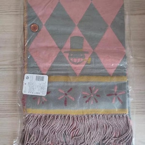 May include: A gray and pink scarf with a diamond pattern and a cartoon character design. The scarf has a fringe at the bottom and a button closure. The scarf is packaged in a clear plastic bag.