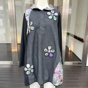 以下が含まれることがあります： ダークグレーのデニム風センターパネル、3つボタンのプラケット、尖った襟が特徴の長袖チュニック。紫、緑、ピンクの色合いの花柄の布製パッチが、花のような形で衣服のアクセントとしてあしらわれています。