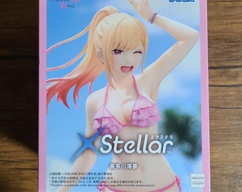 May include: A pink box featuring a Stellar figure of a woman in a pink bikini. The figure has blonde hair and is winking. The box has Japanese text and the SEGA logo. The background is a blue sky.