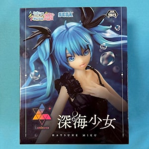 Peut inclure: Un personnage d'anime aux cheveux bleus avec des nœuds noirs dans les cheveux et une robe noire. Le personnage est entouré de bulles bleues. La boîte porte le texte "Hatsune Miku" et "Luminous".