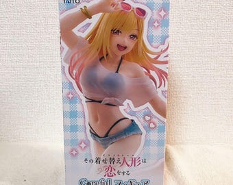 May include: A box featuring a detailed anime figure of a woman in a swimsuit, sunglasses, and a light pink top. The box has Japanese text and the TAITO logo. The background is light blue with star and diamond patterns.