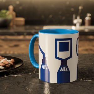 May include: White ceramic mug with a blue interior and handle. The mug features a repeating pattern of blue geometric shapes. The mug is sitting on a black countertop.
