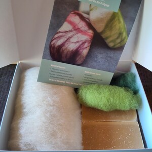 Puede incluir: Un kit de fieltro de jabón DIY en una caja blanca. El kit incluye lana merino natural, lana de color, jabones artesanales e instrucciones. El embalaje presenta ejemplos de jabones afieltrados en rojo, verde y blanco.