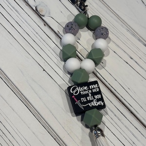 May include: A keychain featuring a loop of sage green, white, and speckled grey beads, a black square bead with the white text "Give me TOUCH HER & I'LL KILL YOU vibes" and a small sword graphic, finished with a silver heart clasp and white tassel.
