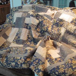 May include: A patchwork quilt draped over a sofa. The quilt features a mix of floral, checkered, and solid fabric squares in shades of blue, beige, and brown. The quilt's design is a mix of patterns and textures, creating a cozy and inviting look.