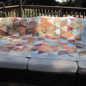 May include: A quilt with a geometric pattern of triangles in shades of brown, orange, pink, blue, and green. The quilt is made of fabric with a mottled, textured appearance. It is displayed on a light brown couch.