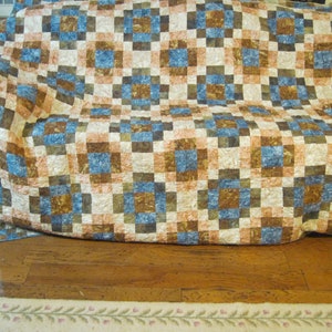 May include: A brown, blue, and white patchwork quilt with a checkered pattern. The quilt is folded and placed on a couch.