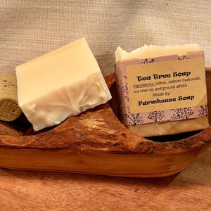 May include: Two bars of handmade soap in a wooden bowl. One bar is white and the other is labeled "Tea Tree Soap" with the ingredients listed. The label also says "Farmhouse Soap".