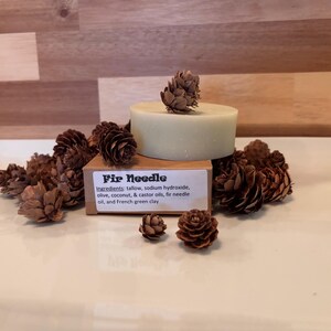 Può includere: Una saponetta rotonda verde pallido con una piccola pigna sopra, circondata da diverse pigne marroni. Un'etichetta recita "Fir Needle" con gli ingredienti. Il sapone è su un piatto bianco.
