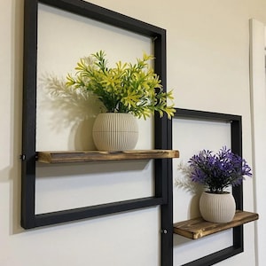 Op de afbeelding: Twee zwarte metalen wandplanken met houten planken. De bovenste plank heeft een plant in een pot met gele bloemen. De onderste plank heeft een plant in een pot met paarse bloemen.