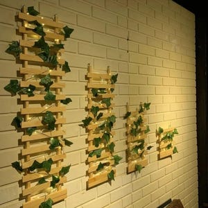 May include: Four wooden wall shelves with a vine of green leaves and white lights. The shelves are arranged in a row on a white brick wall.